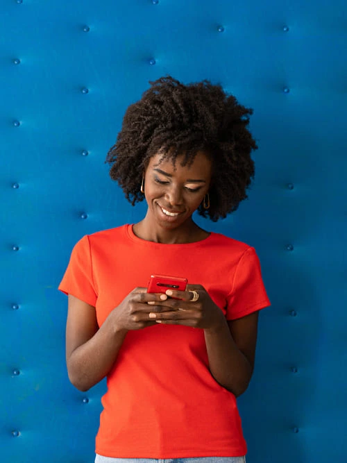 Pessoa com camiseta vermelha, segurando e usando um smartphone vermelho, em frente a uma parede azul acolchoada